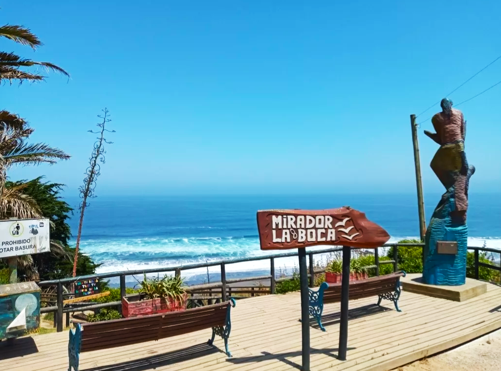 Fotografía del mirador, se aprecia un letrero con su nombre, una estatua y el borde del mar